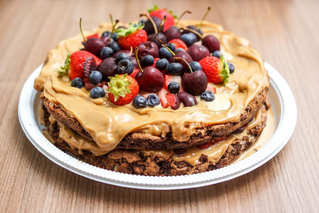 gâteau élégant et appétissant, posé sur une surface en bois clair. Le gâteau est recouvert d'un glaçage lisse et brillant, probablement à base de crème ou de fondant, et décoré de fruits frais comme des fraises et des framboises, ajoutant une touche de couleur vive. La texture moelleuse du gâteau est visible sur une tranche coupée, dévoilant des couches généreuses. L'éclairage naturel met en valeur les détails du dessert, créant une ambiance gourmande et chaleureuse, idéale pour une occasion spéciale ou un moment de partage.






